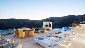A rooftop terrace with a pool, lounge chairs, a dining area, and a view of the sea and hills.