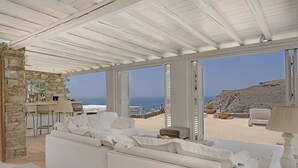 En interiör med vita väggar, en vägg av sten, en vit soffa och utsikt över havet.