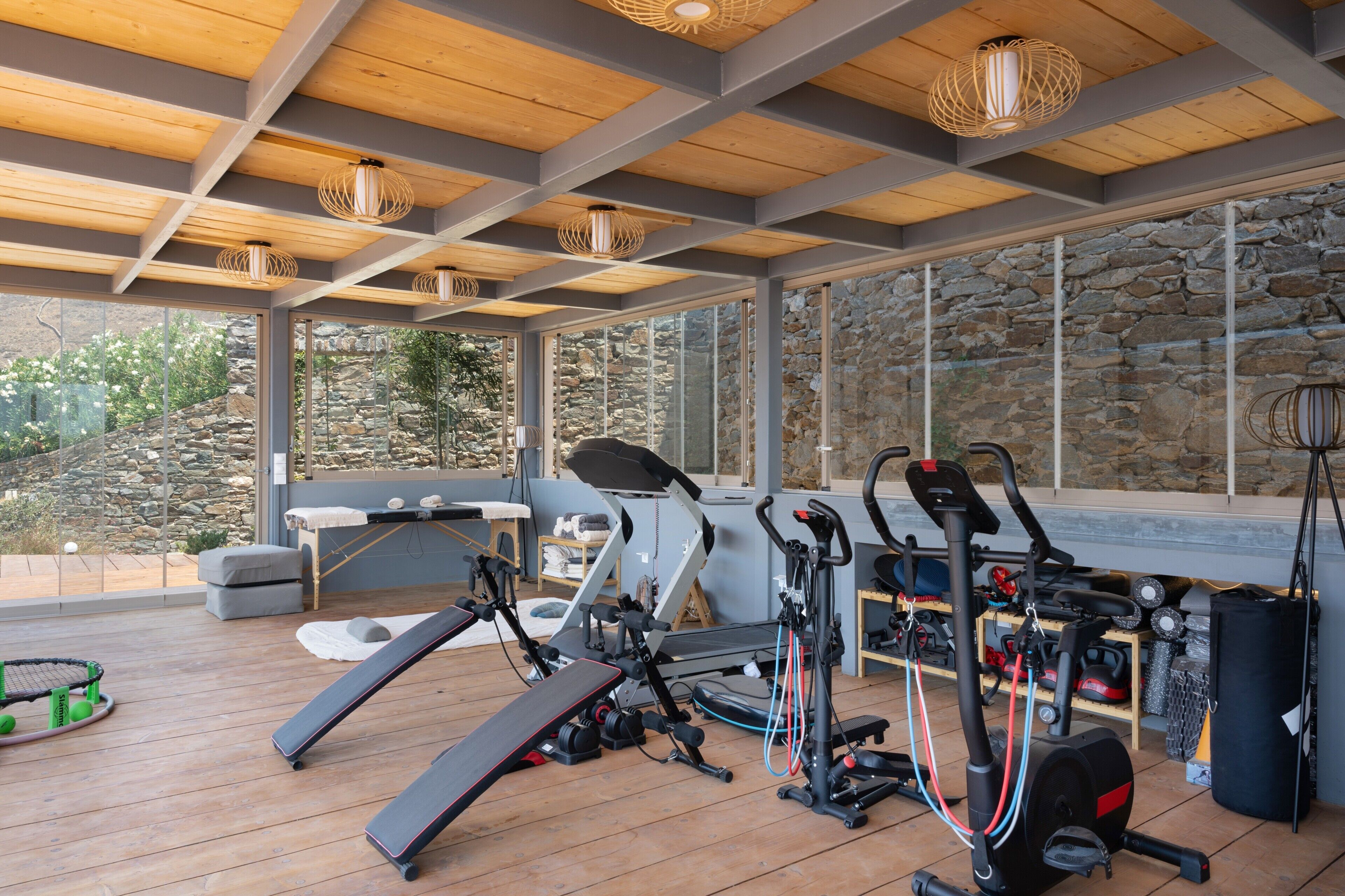 A gym with exercise equipment, large windows, and a stone wall.
