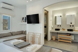 Habitación de hotel moderna con sofá, televisor, baño con espejo y lavamanos, y vista al exterior.