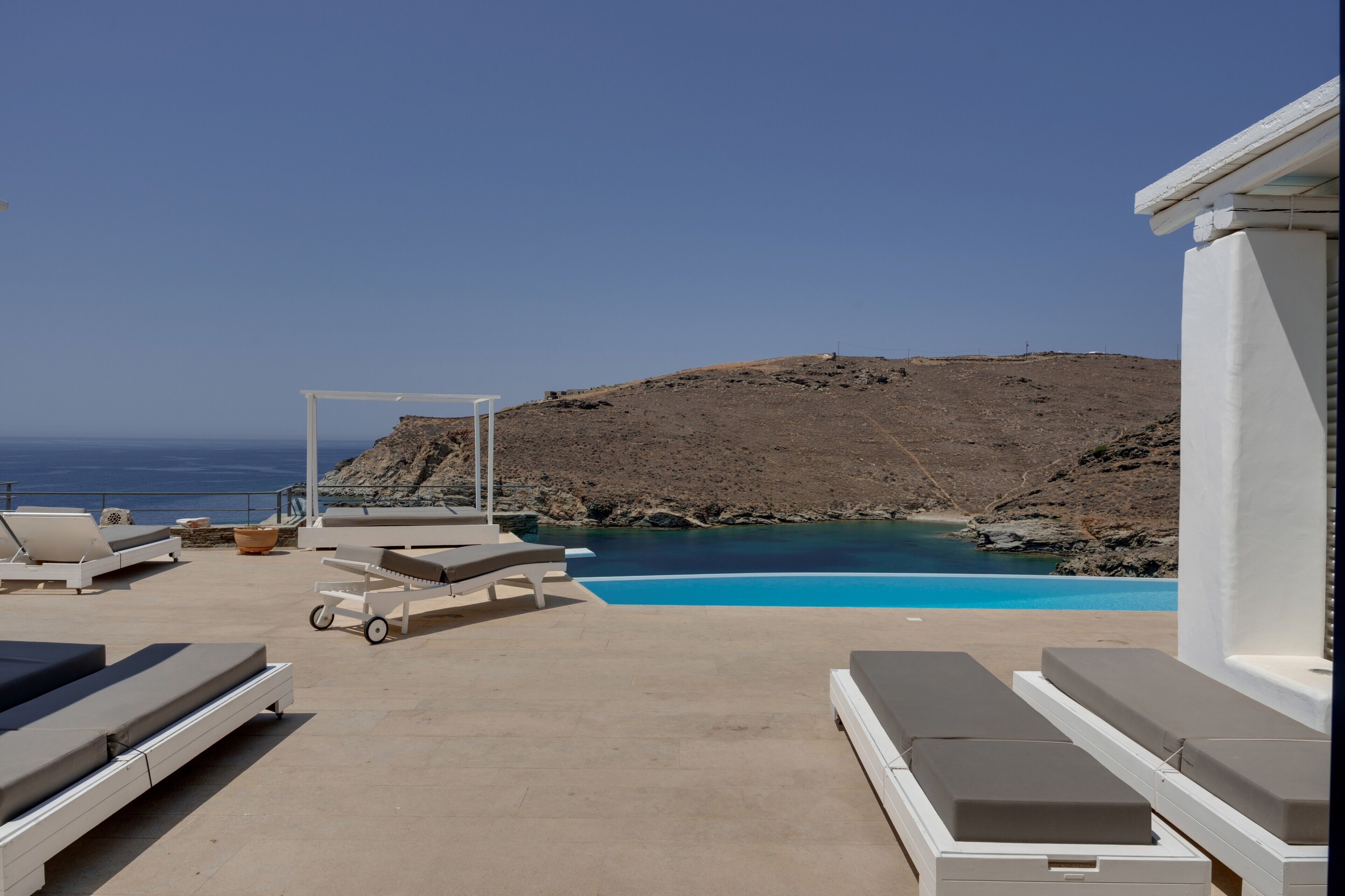 A poolside area with lounge chairs, a small table, and a view of the sea and rocky terrain.