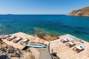 Un muelle de madera con sillas de descanso y una tabla de surf, con vista a un mar azul cristalino y una costa rocosa.
