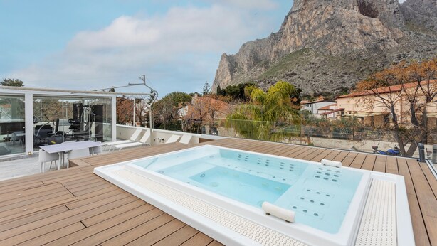 A wooden deck with a hot tub, a glass-enclosed area with outdoor furniture, and a mountain backdrop.