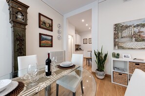 A dining area with a glass table, chairs, a clock, framed pictures, and a potted plant.