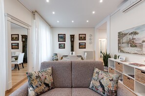 A living room with a sofa, cushions, a dining area with chairs, a table, and wall-mounted artwork.