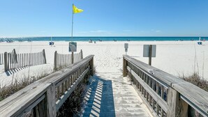 En træbro fører til en sandstrand, med et gult flag, en klar blå himmel og et roligt hav.