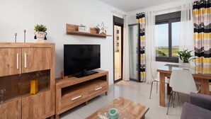 A modern living room with a wooden TV stand, a flat-screen TV, a dining table with chairs, and a view of the outdoors through a large window. - Playa Marinsa Beach – Pool, Padel, and Beach in Torre del Mar (Torre del Mar)
