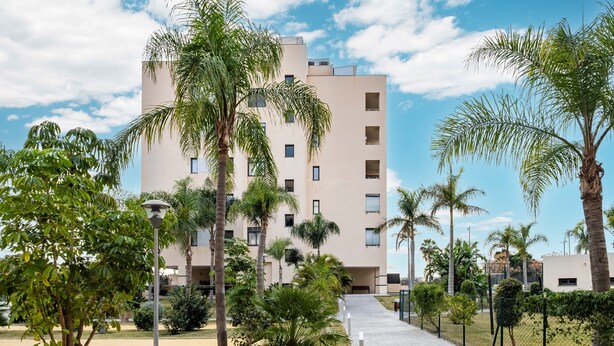 A modern building with multiple floors, surrounded by palm trees and a paved walkway. - Playa Marinsa Beach – Pool, Padel, and Beach in Torre del Mar (Torre del Mar)