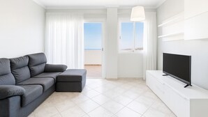 Un salon moderne avec un canapé gris, un téléviseur à écran plat, des murs blancs et un balcon offrant une vue dégagée.