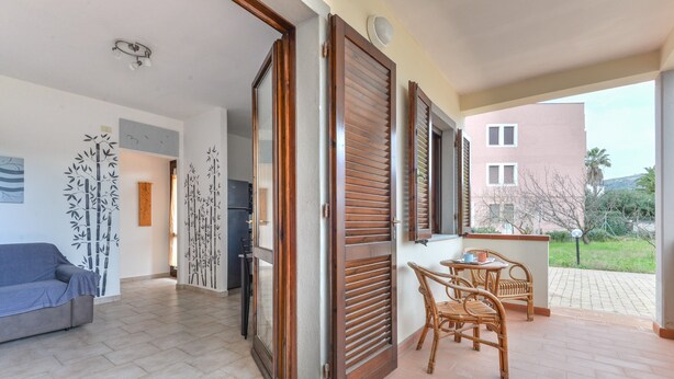 Un salon avec un canapé, un coin repas avec des chaises et une table, un espace cuisine, et une vue sur un bâtiment et la verdure environnante.