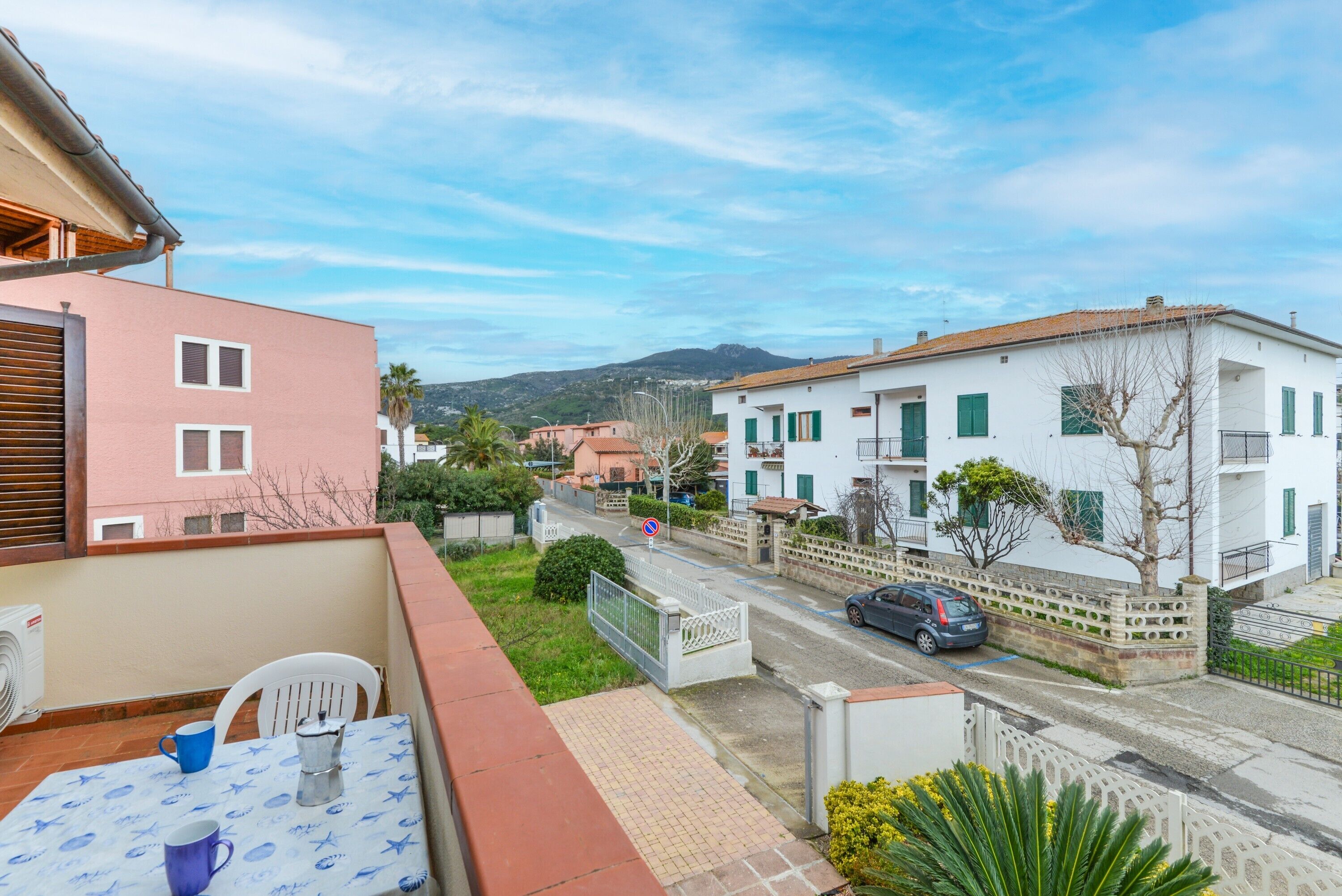 Un balcon avec une table et des chaises, une vue sur la rue avec des immeubles et une voiture, et un ciel dégagé.