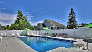 A rectangular swimming pool with a metal ladder, surrounded by white lounge chairs and a green umbrella.