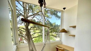 Une pièce dotée d’une grande fenêtre, d’un fauteuil suspendu, d’un banc en bois et d’une étagère fixée au mur.