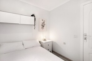 Une chambre à coucher avec un lit, une table de chevet avec une lampe, un miroir fixé au mur, un tableau encadré et une porte.