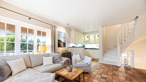 Un salon spacieux avec un grand canapé, une table basse et un fauteuil. Au fond, on aperçoit une cuisine avec des meubles blancs et un escalier menant à l’étage supérieur.