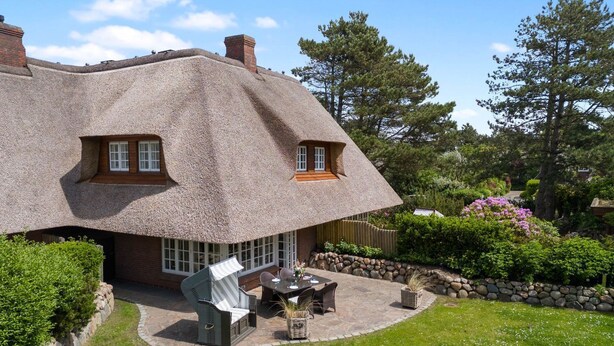 Une maison avec un toit de chaume, une terrasse avec des meubles d’extérieur et un jardin bien entretenu.