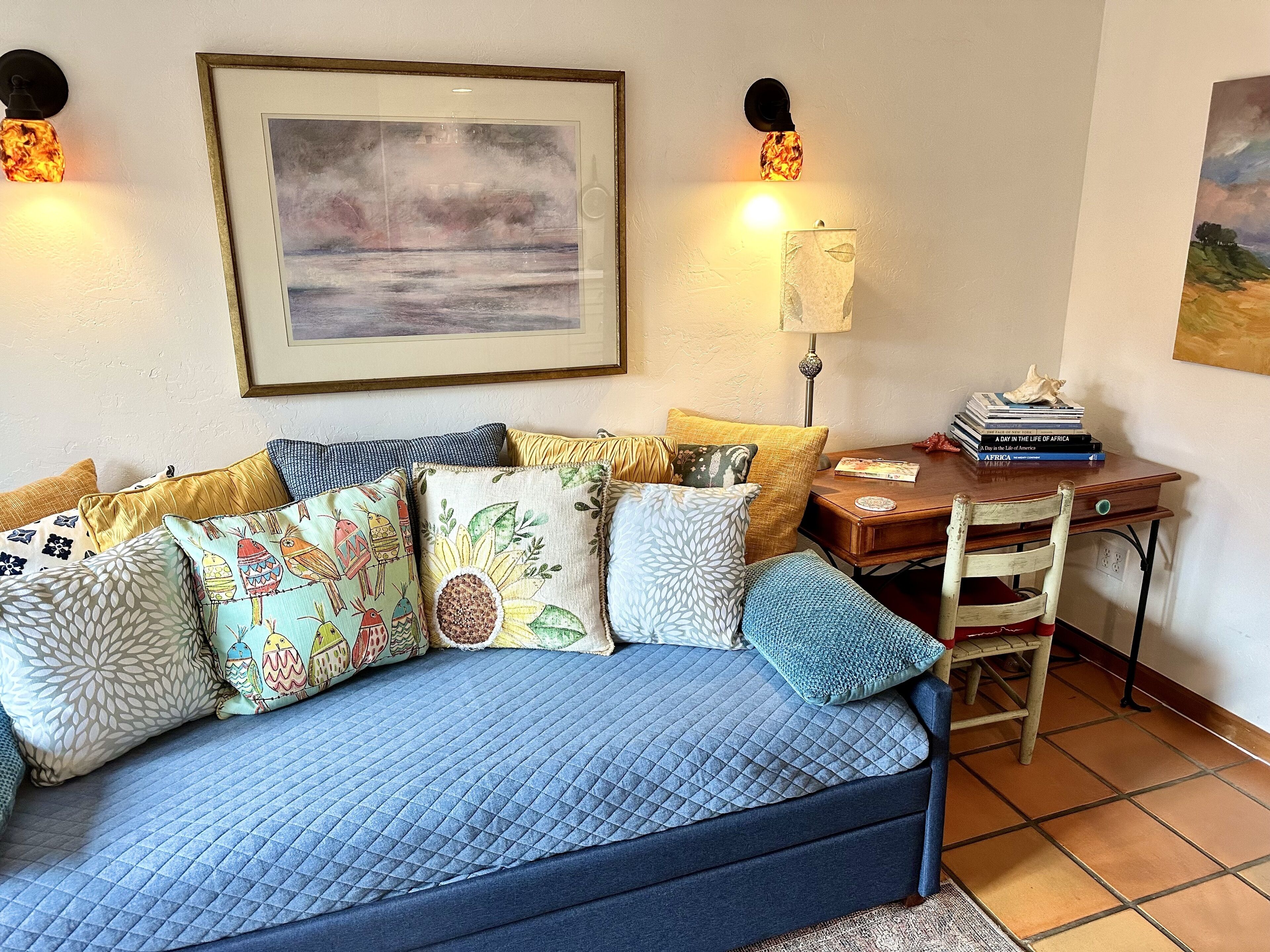 Una sala de estar acogedora con un sofá azul, cojines coloridos, un cuadro enmarcado y un escritorio de madera con libros.