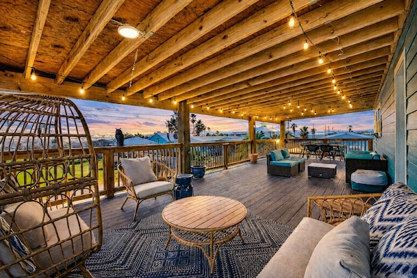 A covered outdoor deck with wooden beams, string lights, wicker furniture, and a view of the landscape.