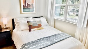A neatly made bed with white bedding and decorative pillows, a framed cityscape poster on the wall, a bedside lamp, and a window with curtains. - Minutes from downtown Austin! Cozy bungalow w/ fire pit; perfect for families! (Austin)