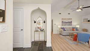 A modern living room with a grey sofa, a coffee table, and a blue armchair. There is a white door with a mirror and wall sconces on the left. - Minutes from downtown Austin! Cozy bungalow w/ fire pit; perfect for families! (Austin)