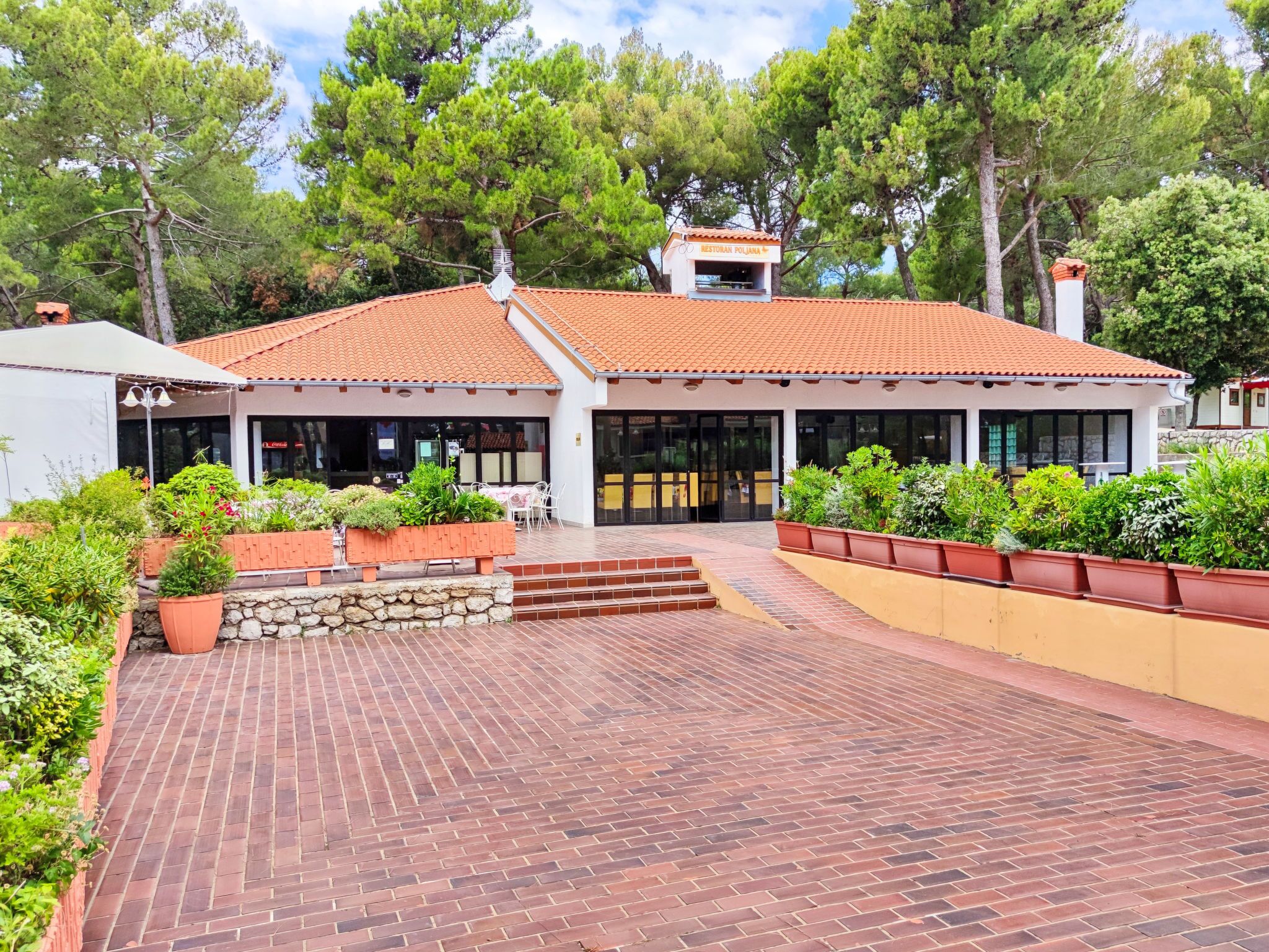A building with a tiled roof, a paved entrance area, and a garden with potted plants.