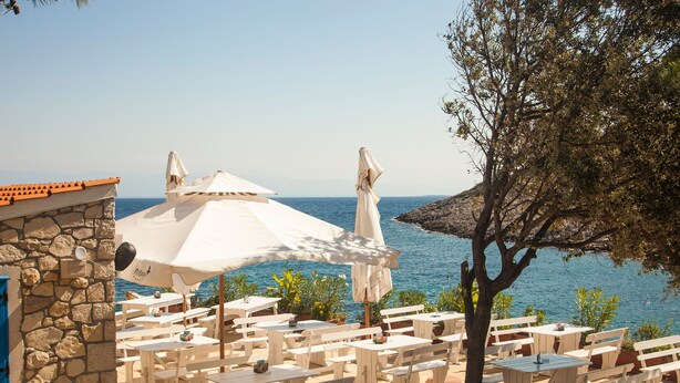 Un espace extérieur aménagé avec des tables et des bancs blancs, des murs en pierre et des parasols, offrant une vue sur la mer.