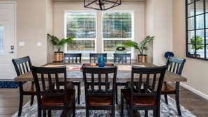 A dining area with a wooden table, six chairs, and a chandelier. There are potted plants on the table and windows with blinds. - Mountain View House with hot tub, putting green, stunning views! (Oakhurst)