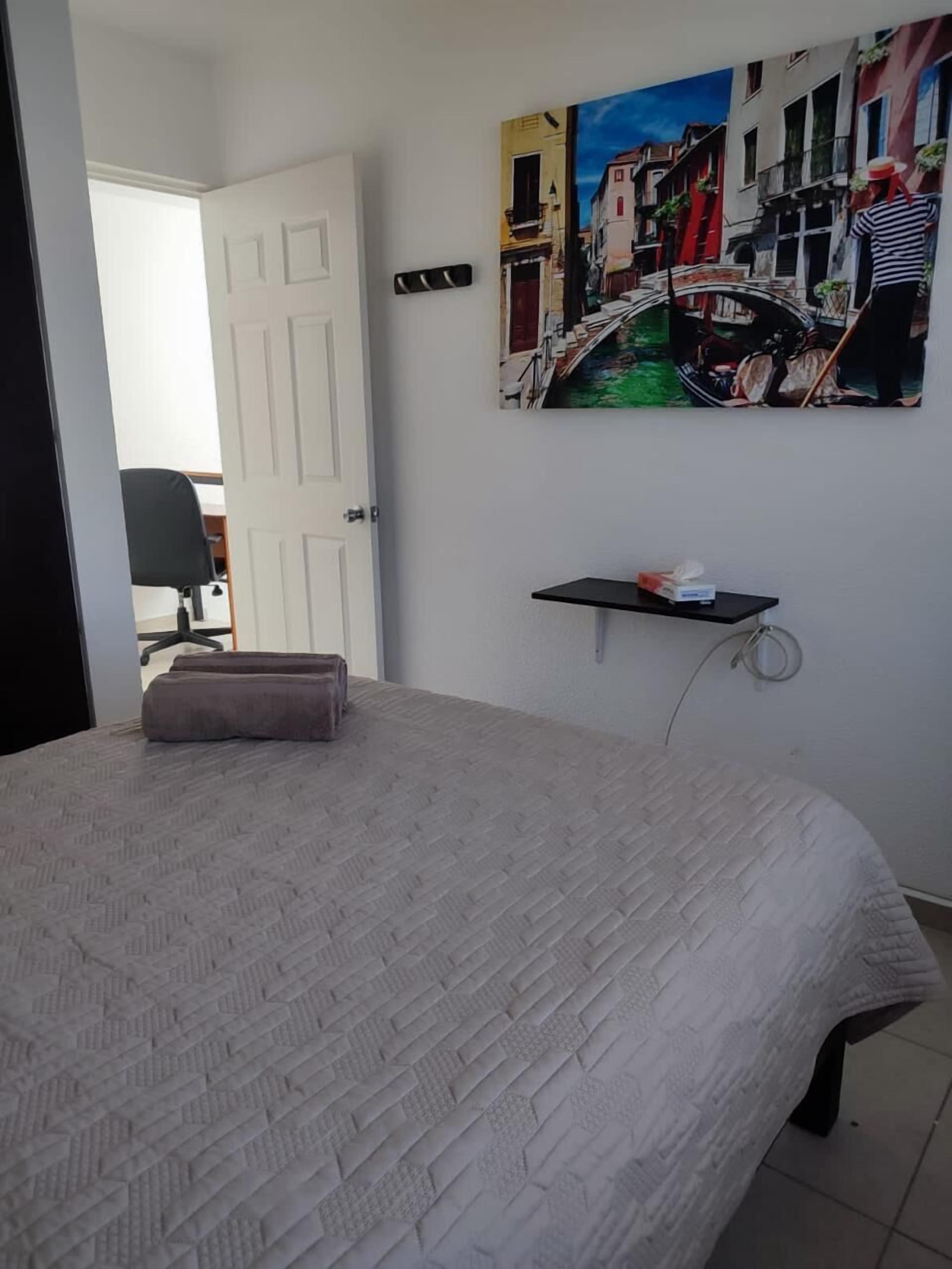 Un dormitorio con cama, puerta, silla, estante en la pared y un cuadro con una escena de canal.
