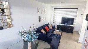 A modern living room with a glass dining table, dark blue sofa, and a TV in the background. - CASA LAUREL (Corregidora)