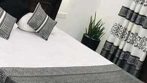 A neatly made bed with a geometric-patterned bedspread and pillows, a dark wooden headboard, a wall-mounted light fixture, and a potted plant beside the bed. - Ibeeza Motel (Batigama)