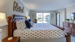Una habitación de hotel con una cama grande, un cabecero de madera, vista al agua, un televisor y un cartel con la palabra 'Blessed'.