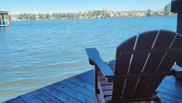 Eine Holzterrasse mit einem Holzstuhl, der auf einen ruhigen See ausgerichtet ist, umgeben von Häusern und Bäumen.