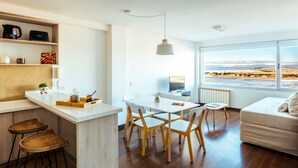 A modern kitchen with a white island, wooden stools, and a dining area with chairs. A large window offers a view of a landscape. - Moat Apartamento (Ushuaia)
