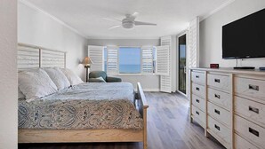 A bedroom with a bed, a TV, a dresser, and a window with shutters. - Tiffany Plaza in Longboat Key, FL (Longboat Key)