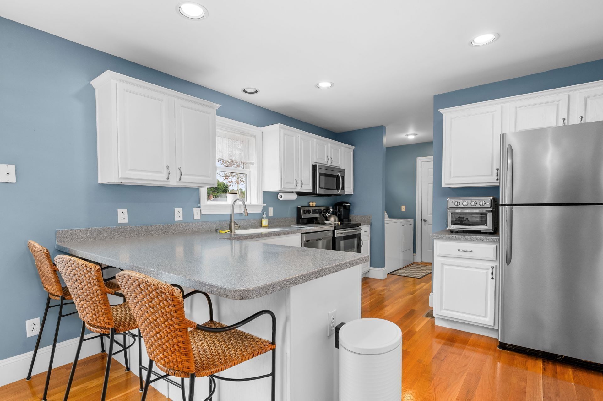 A modern kitchen with white cabinets, a stainless steel refrigerator, a microwave, and a built-in oven. There are two wicker chairs at the island and a trash can nearby.
