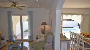 Una sala de estar con un sofá, una mesa de centro, una mesa de comedor con sillas y vista a una piscina a través de una puerta abierta.