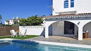Una piscina con zona de patio, mobiliario exterior y un edificio blanco con arcos.