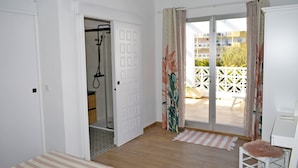 Una habitación con piso de madera, una cama con colcha a rayas, un sillón con cojín mullido, un ventanal corredero que da a un área exterior con cerca blanca y un equipo de aire acondicionado en la pared.