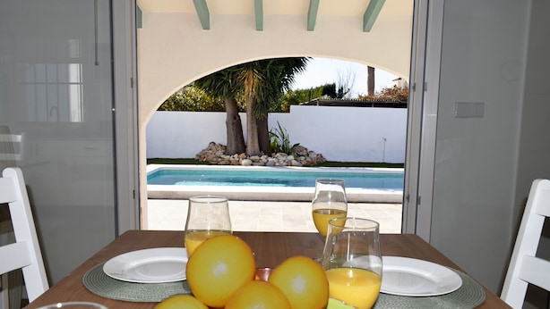 Una mesa de comedor con frutero, vasos y platos, con vistas a una piscina y palmeras.