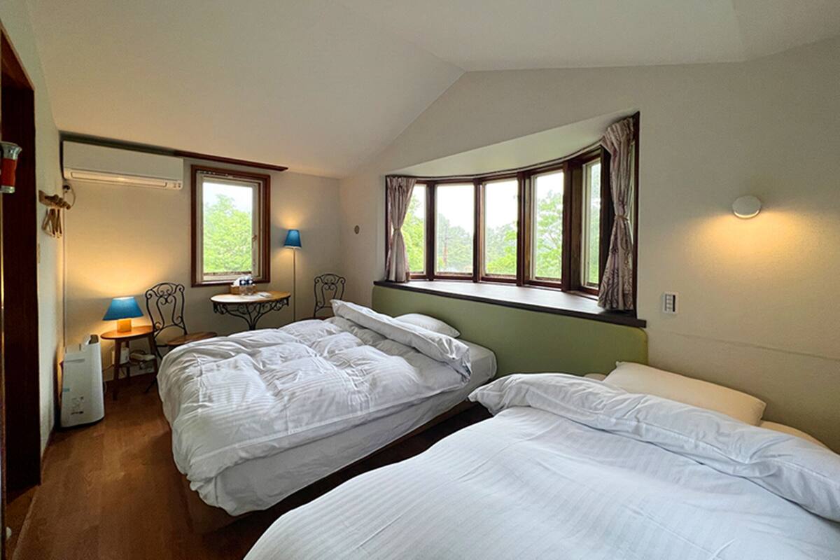 A room with two single beds, a small table with a chair, a window with curtains, and a lamp.