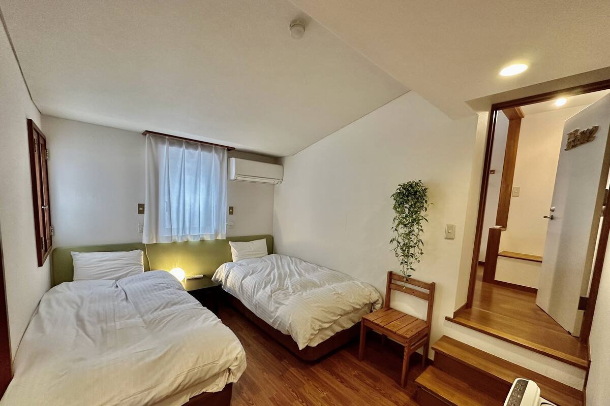 A hotel room with two single beds, a small table, a chair, a potted plant, a mirror, and a wooden floor.