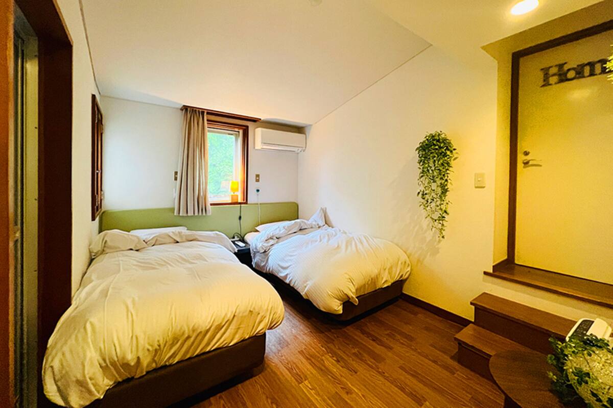 A room with two beds, a window with curtains, an air conditioning unit, a plant, and a door labeled "Home".