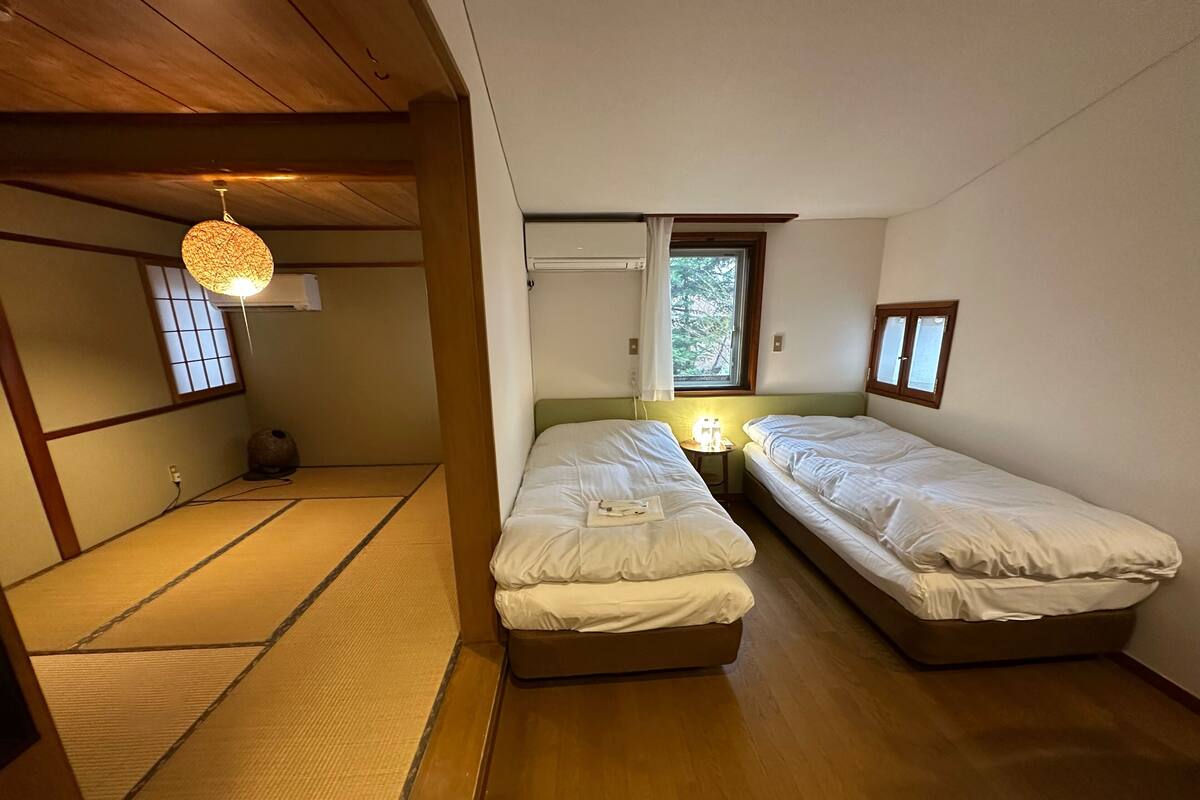 A room with two single beds, a small table with a lamp, a window with a view of trees, and a traditional tatami mat floor.