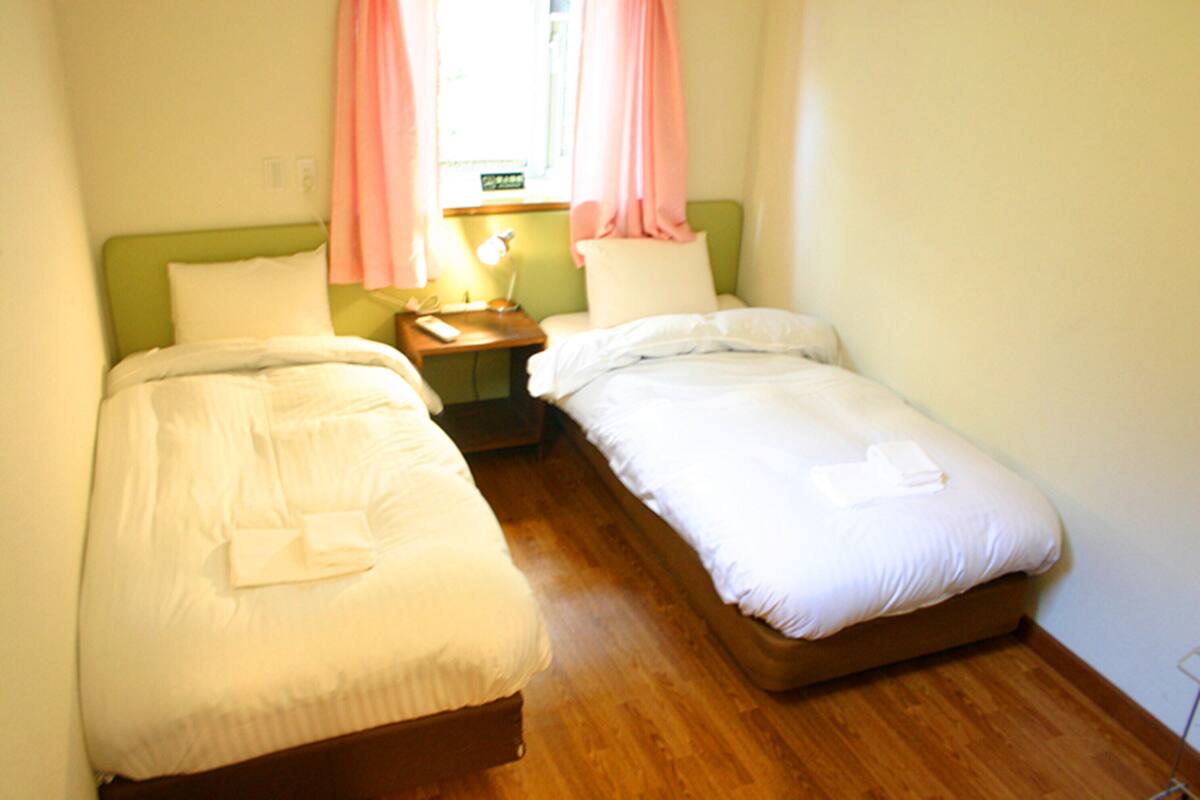 A small, clean room with two single beds, a bedside table with a lamp, and a window with pink curtains.