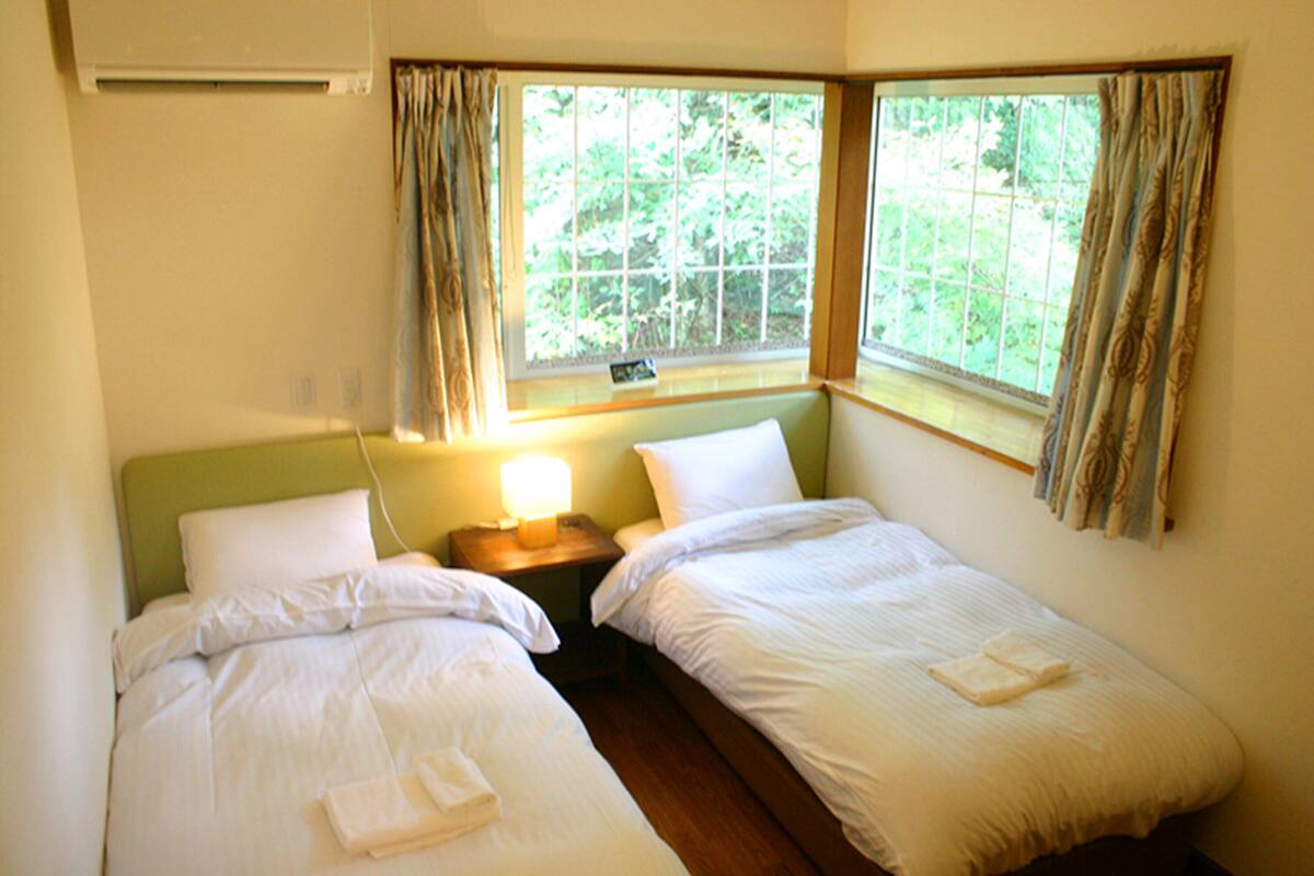 A twin room with two beds, a nightstand, a window with curtains, and a wall-mounted air conditioner.