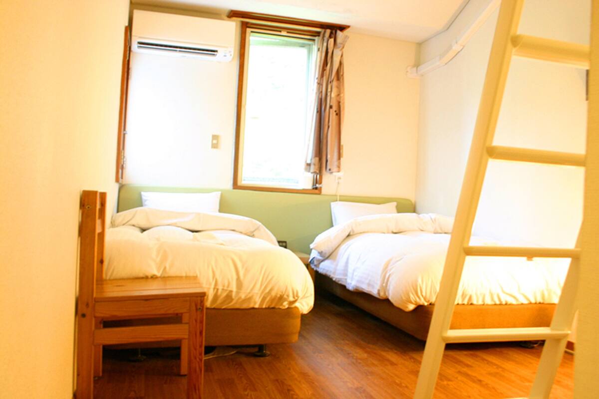 A room with two beds, a wooden chair, a ladder, and a window with curtains.