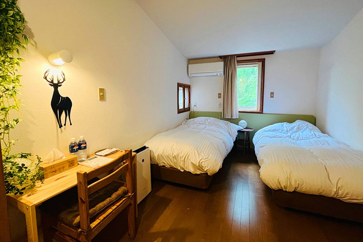 A small, well-lit room with two beds, a desk, and a chair. There is a wall-mounted lamp, a window with curtains, and a plant on the left.