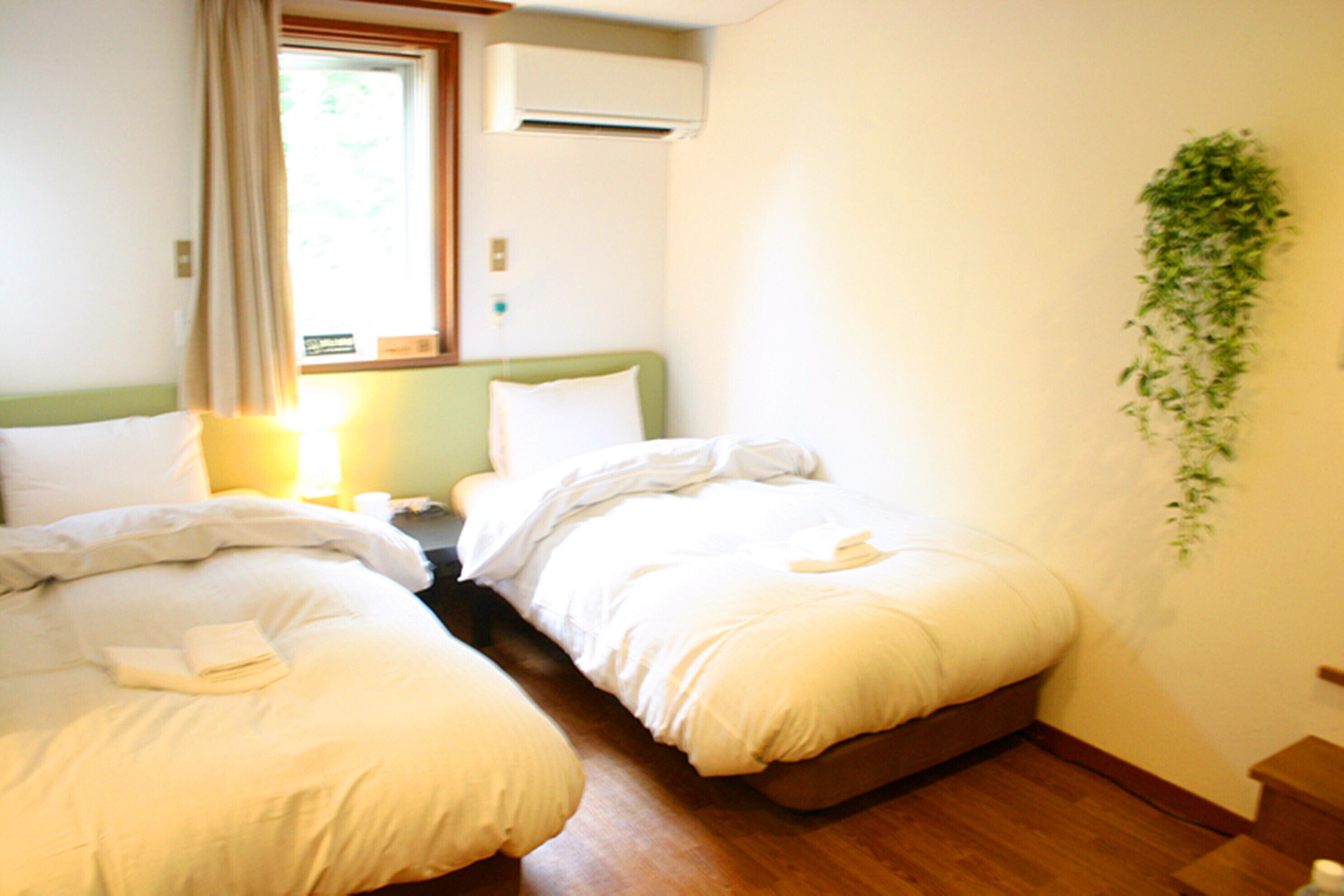 A hotel room with two beds, a window with curtains, an air conditioning unit, and a wall-mounted plant.