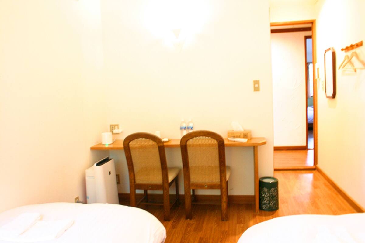 A small hotel room with two single beds, a wooden desk with chairs, a trash bin, and a door leading to another room.