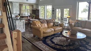 A spacious living room with a large leather sofa, a coffee table, and a patterned rug. There are multiple windows providing natural light and views of the outdoors. -  Secluded Chickalah Mt. retreat overlooking beautiful Harkey Valley! (Belleville)